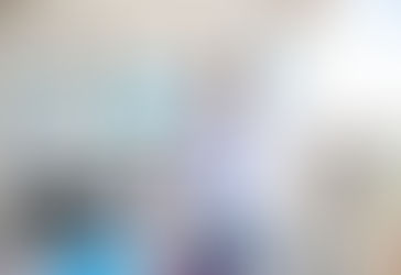 Smiling person in a blue shirt leans on a glass desk with photos on the wall. Bright room with a large window and modern chair.