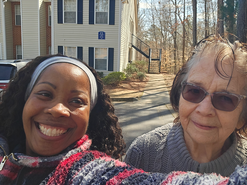 Two women smiling outdoors in front of a building with blue shutters. One wears a colorful jacket, the other a gray sweater. Clear, sunny day.