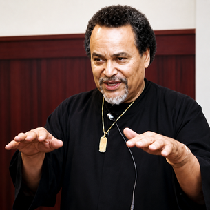 Man in a black shirt and gold necklace speaks with both hands raised in a wood-paneled room, looking focused and animated.