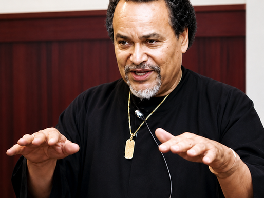 Man in a black shirt and gold necklace speaks with both hands raised in a wood-paneled room, looking focused and animated.