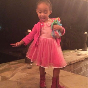 Little girl in a pink dress and boots poses by a stone fireplace, sticking out her tongue and holding a colorful doll.