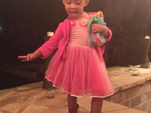 Little girl in a pink dress and boots poses by a stone fireplace, sticking out her tongue and holding a colorful doll.
