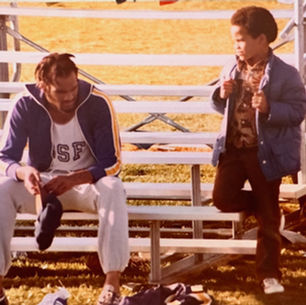 Two boys on sunlit bleachers, one seated looking down while the other stands holding his jacket at a grassy field.