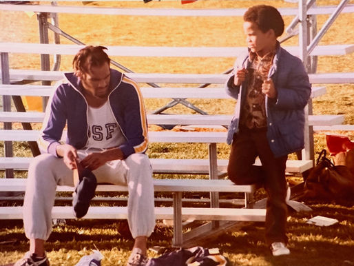 Two boys on sunlit bleachers, one seated looking down while the other stands holding his jacket at a grassy field.