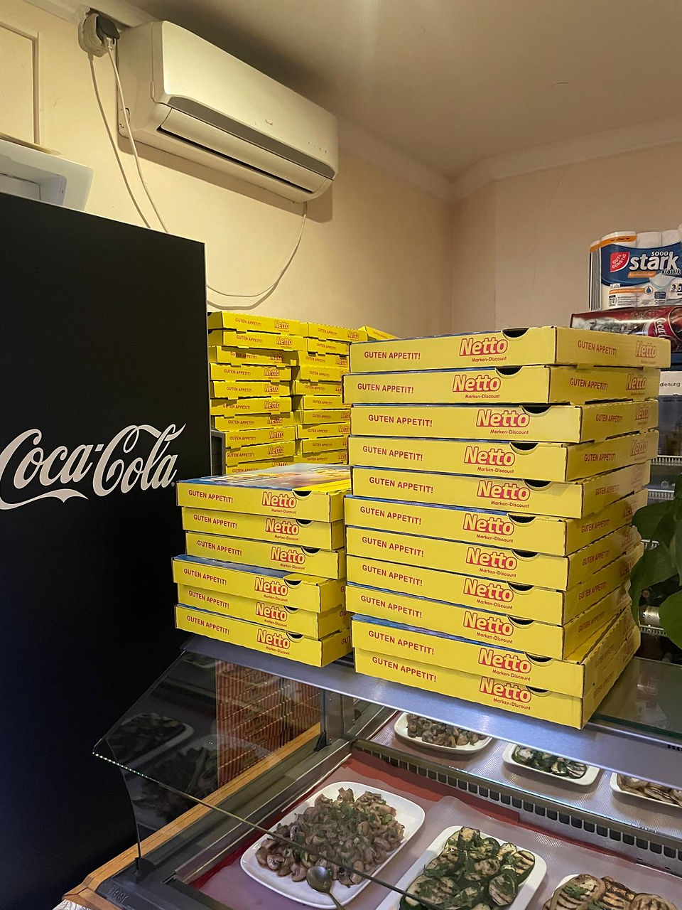 Ein enger Lagerraum mit einem hohen Stapel Netto-Pizzakartons, die an der Wand lehnen. Die gelben Kartons mit Netto-Logo sind für die Verpackung von Pizzen vorbereitet, während im Hintergrund eine Coca-Cola-Werbung an der Wand hängt.