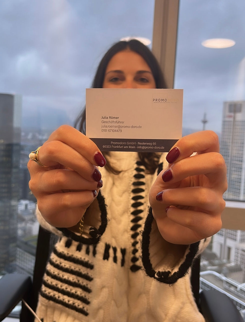 Eine Frau mit dunklen Haaren hält eine Visitenkarte in die Kamera, die den Namen Julia Römer, ihre Position als Geschäftsführerin und Kontaktdaten enthält. Sie trägt einen cremefarbenen Pullover mit schwarzen Mustern, und ihre Nägel sind in einem dunklen Rotton lackiert. Im Hintergrund ist eine moderne Büroumgebung mit großen Fenstern und einer Stadtansicht zu sehen.