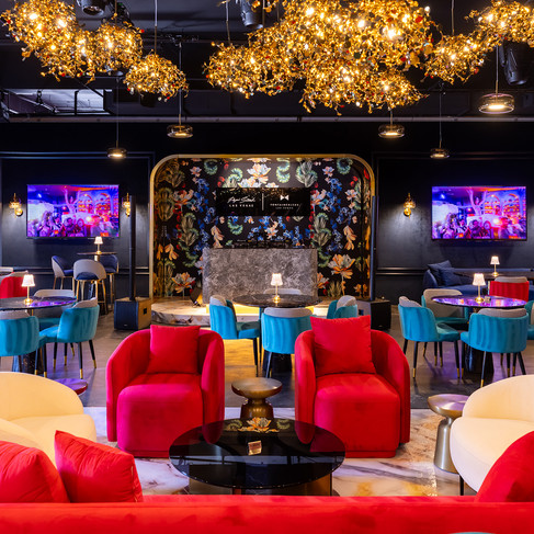 A high-end lounge and dining space at Papi Steak Las Vegas, featuring plush red and ivory seating, black marble tables, and bold floral-patterned walls framed in gold. The ceiling is adorned with dazzling golden chandeliers, casting a warm glow over the elegant setting. Deep blue and teal chairs complement the sophisticated ambiance, while large display screens showcase dynamic event visuals.