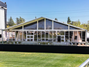 Large glass-walled pavilion on green lawn, sunny day, trees in background. Inside are chairs and tables. "Platinum" sign visible.