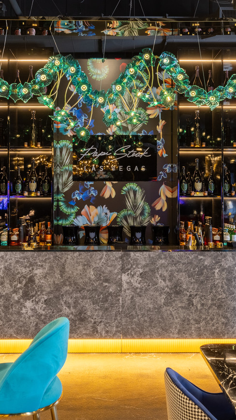A luxurious bar setup at Papi Steak Las Vegas, featuring a sleek dark marble counter with gold accent lighting. The backdrop showcases an illuminated liquor display with floral-patterned decor, while a striking emerald-green chandelier adds a dramatic touch. Plush teal chairs and marble-top tables enhance the upscale ambiance, with two screens displaying event visuals in the background.