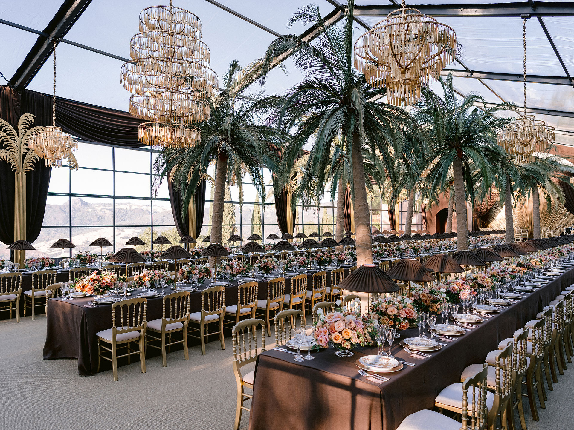 Interior of luxury clear-span tent with chandeliers, palm trees, and long banquet tables set for an elegant event.