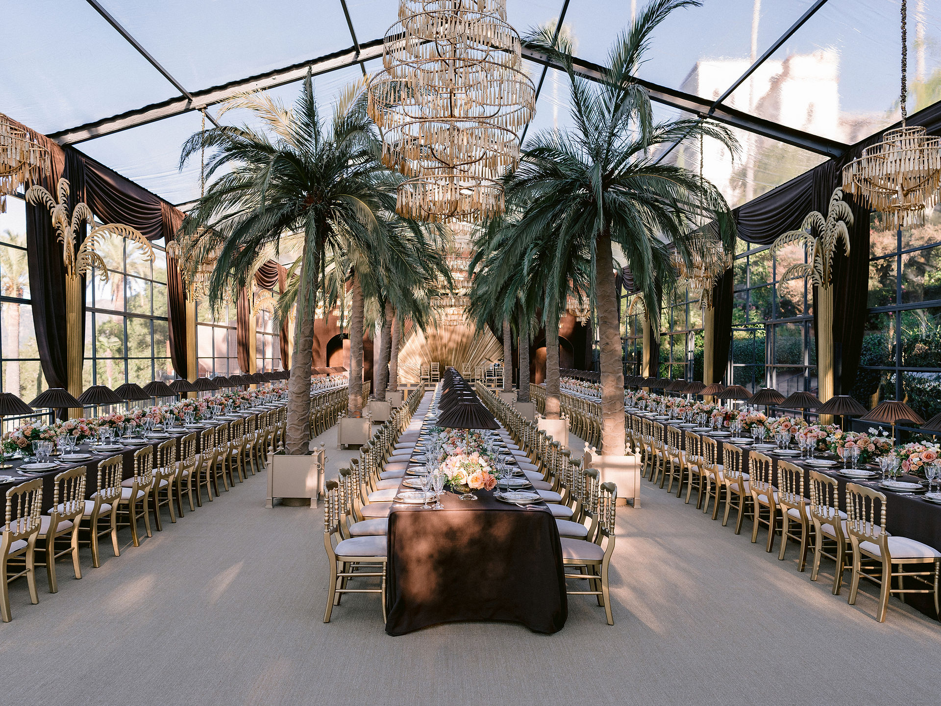 Sunset view inside a clear tent with palm tree décor, gold chairs, chandeliers, and opulent table settings.