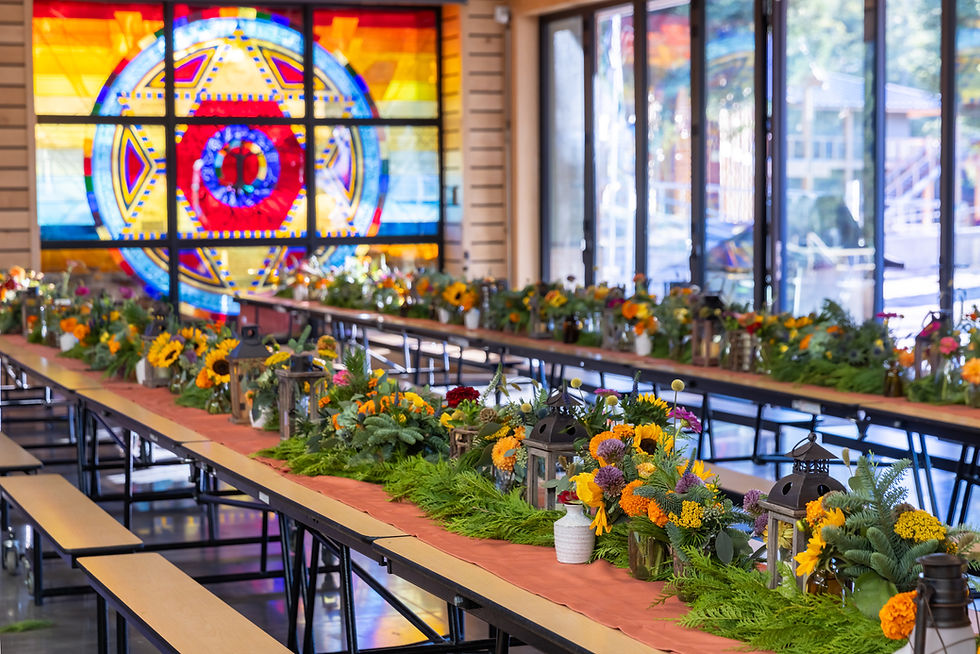 Tables with vibrant flower arrangements and lanterns, set in a hall with a large colorful stained glass window in the background.