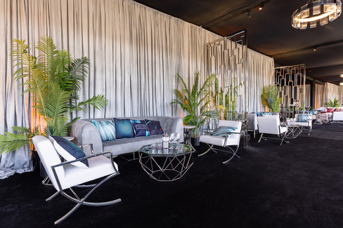 A luxurious event lounge featuring a plush gray sofa, white leather chairs, modern geometric tables, metallic dividers, and soft draping, accented with tropical greenery.