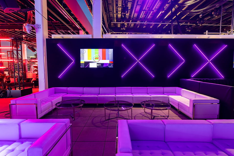 Modern lounge with purple-lit sofas, round tables, and neon lights on a black wall. A colorful screen displays geometric patterns.