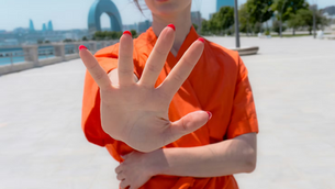 Woman in orange holds up an open hand with red nails on a sunny waterfront promenade, city skyline blurred behind.
