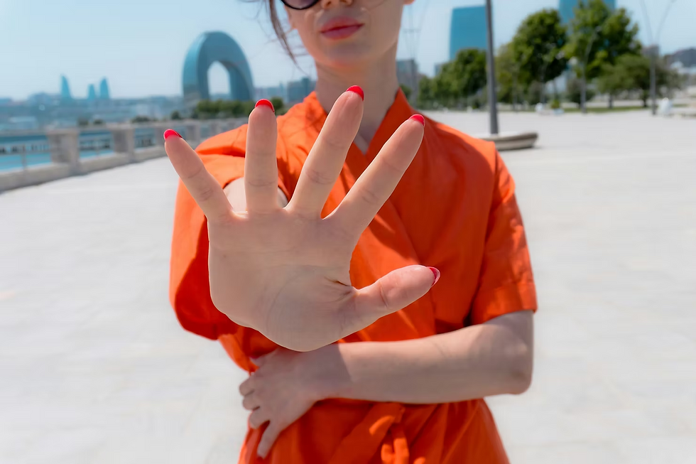 Woman in orange holds up an open hand with red nails on a sunny waterfront promenade, city skyline blurred behind.