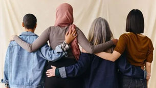 Four people stand with arms around each other from behind against a plain beige backdrop, suggesting friendship and unity.