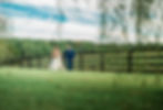 Bride and groom walk hand in hand in a grassy field with a wooden fence and string lights. The sky is partly cloudy, creating a serene mood at Oak Creek Farm.