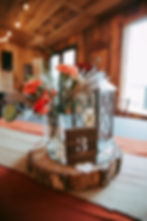 Floral centerpiece with orange flowers, lantern, and table number 3 on a wood slice. Rustic indoor setting with soft lighting at Oak Creek Farm.
