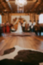 Bride and groom hold hands under floral arch in rustic barn wedding. Bridesmaids in red dresses, groomsmen in hats. Warm lighting at Oak Creek Farm