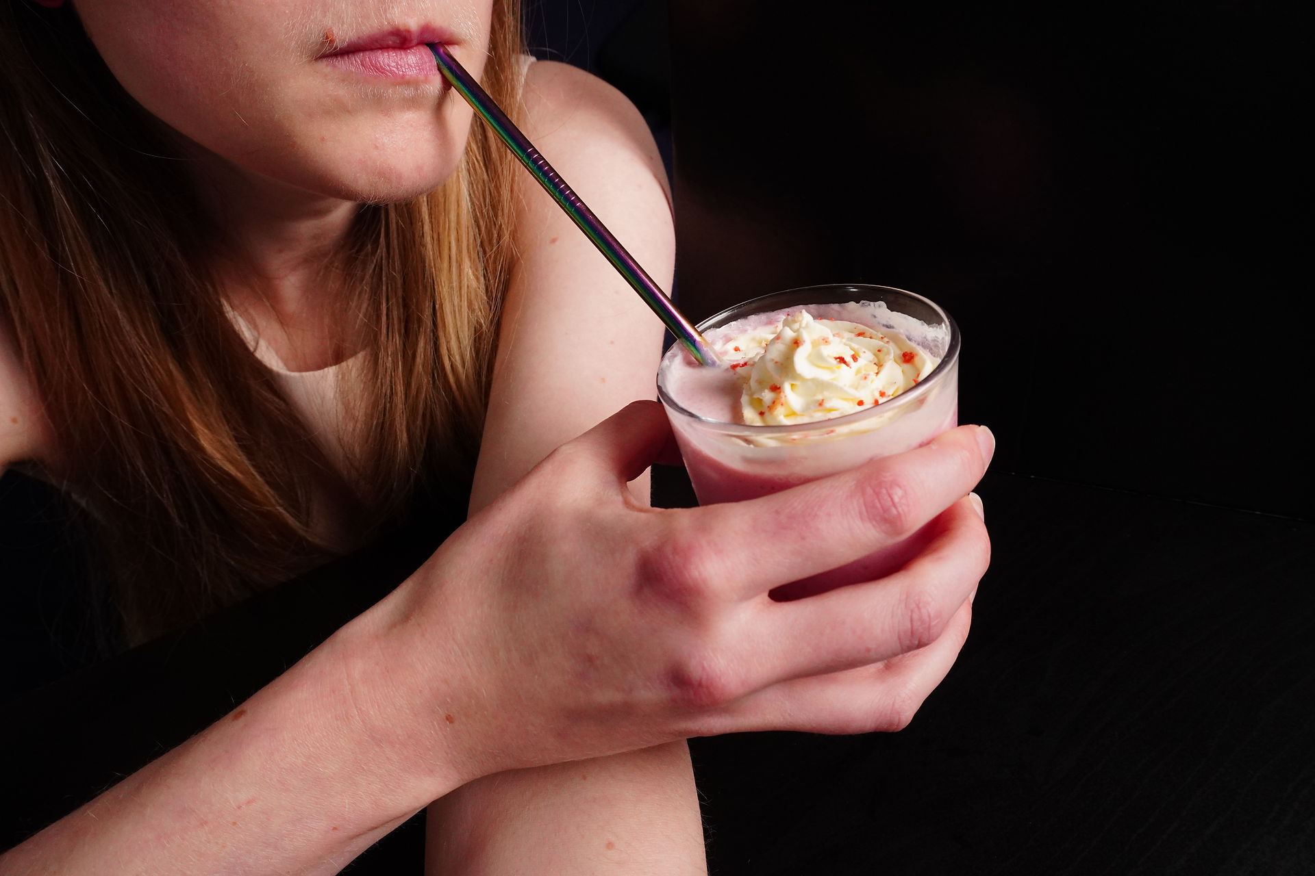 blonde girl drinking pink drink with cream on top, metal straw, black background