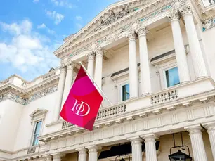 Institute of Directors — 116 Pall Mall. A Neoclassical building with white columns and ornate details. A red flag with "IoD" text waves in front, set against a blue sky.