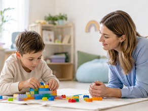 Therapist watches smiling as boy builds colourful block tower on the floor in a warm clinic space with rainbow wall art.