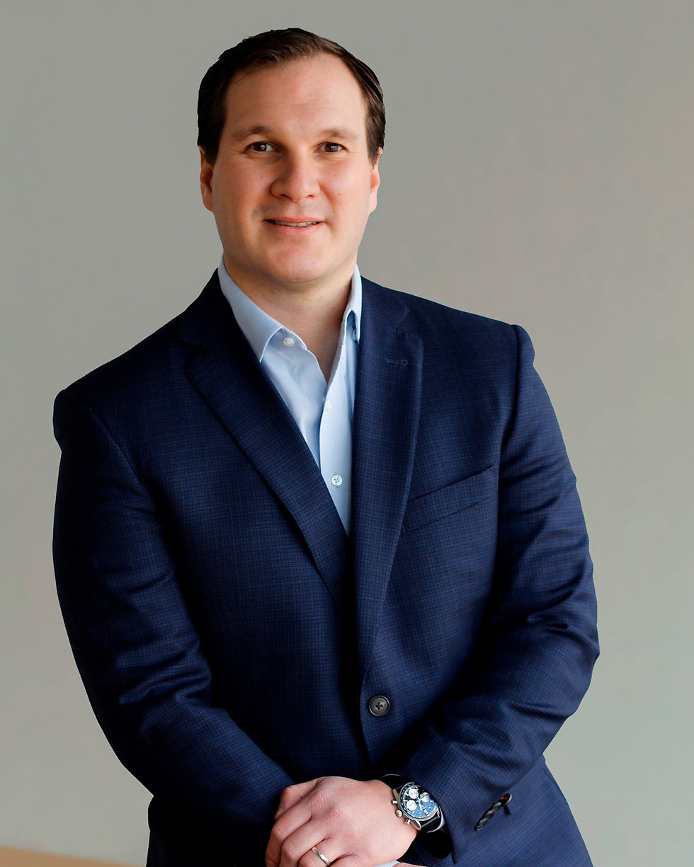 Hombre con traje azul marino y camisa celeste, posando con leve sonrisa sobre fondo gris.