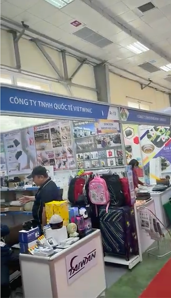 Trade show booth with luggage and electronics display in Vietnam International Trade Fair. Visitors interact. Blue sign reads "CÔNG TY TNHH QUỐC TẾ VIETWNIC." Bright, busy atmosphere.