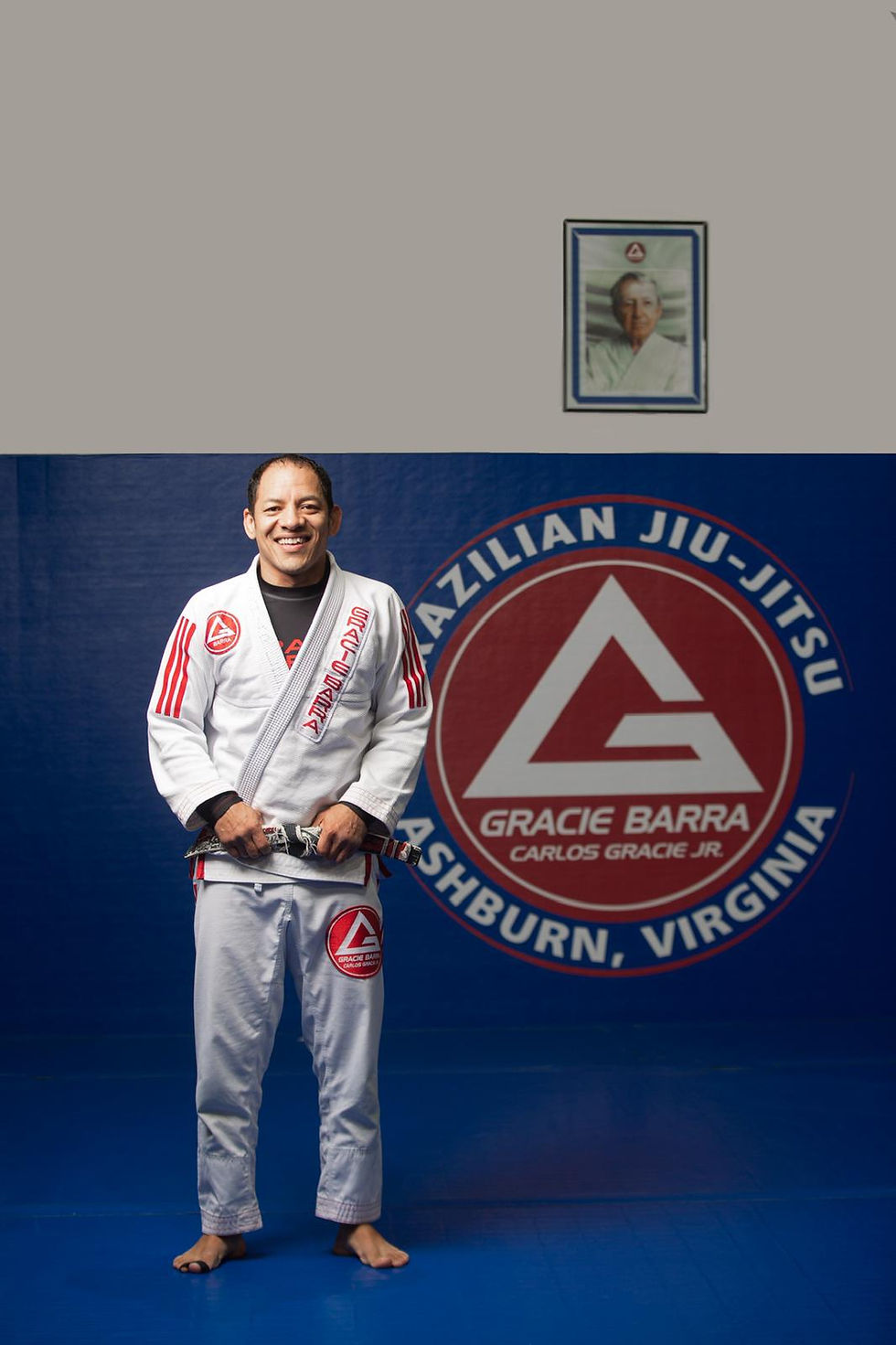 Professor Charles from the Gracie Barra Ashburn Jiu-Jitsu academy, with short dark hair, wearing a white gi with red details and a black jiu-jitsu belt, is standing on a blue mat in Virginia.