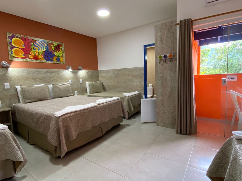 Suíte quíntupla da Pousada Kaliman com uma cama de casal e duas de solteiro, enxoval bege, decoração moderna com parede em tom laranja, frigobar e sacada com vista parcial a praia de Ubatuba