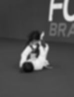Two children, likely girls, practicing jiu-jitsu on a dark mat. One child is lying on her back wearing a white gi. The other child is sitting on top with her legs over, also wearing a gi.