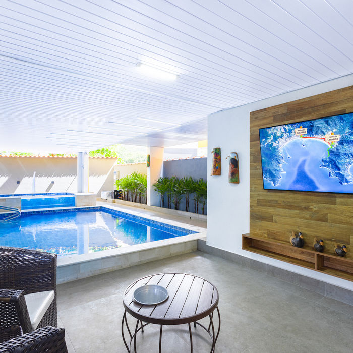 Piscina retangular azul com hidromassagem e borda clara em área de lazer interna parcialmente coberta e iluminada. TV exibe praia sobre painel de madeira com prateleiras. Área de estar relaxante com mesa redonda e duas cadeiras de vime ao lado da piscina com piso de concreto cinza e vasos de plantas retangulares em Ubatuba na Pousada Kaliman