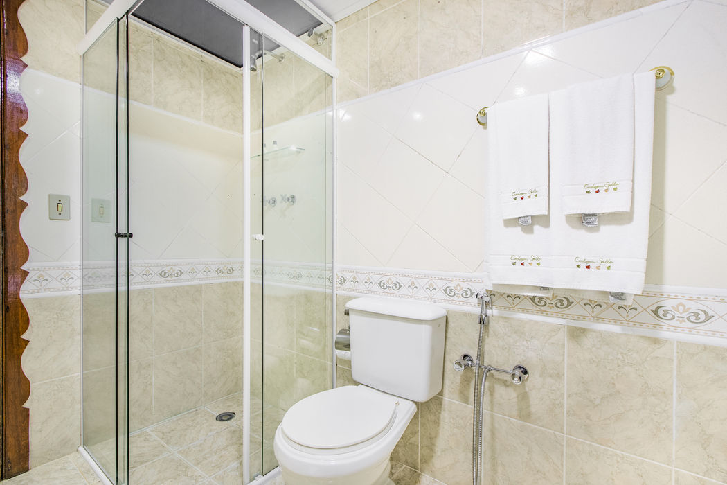 Banheiro pequeno com azulejos bege claros e piso de cerâmica branco, chuveiro com porta de vidro, vaso sanitário e toalhas brancas penduradas na Estalagem Spiller em Monte Verde