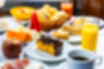 Café da manhã variado em Ubatuba com um pedaço de bolo de cenoura coberto com chocolate em destaque. Frutas frescas como melancia, mamão e maçã, além de sanduíches e pães em uma cesta, também estão na mesa na Pousada Kaliman