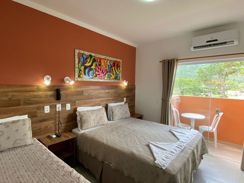 Acomodação em Ubatuba com cama de casal, cama de solteiro e pintura marinha colorida na parede cor de ferrugem. Varanda com mesa e cadeiras brancas, vista para área verde e ar-condicionado instalado. Paredes brancas, cabeceira de madeira clara com arandelas e mesas de cabeceira escuras na Pousada Kaliman
