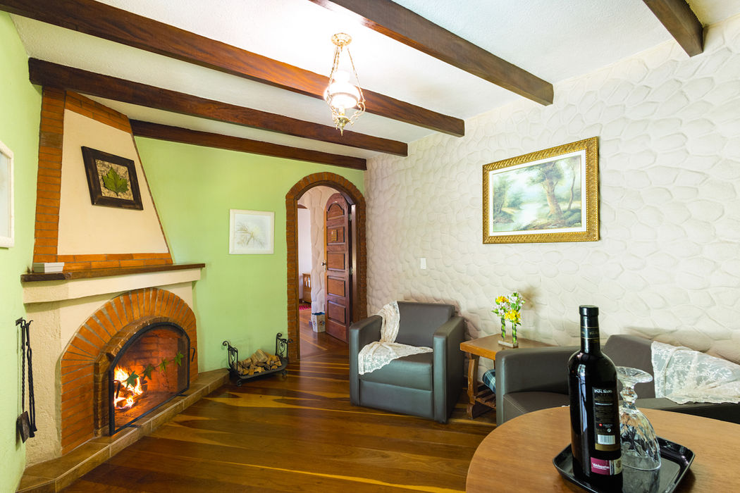 Sala de estar aconchegante e rústica na Estalagem Spiller, Monte Verde, com lareira acesa. Piso de madeira, teto com vigas escuras expostas, uma parede texturizada branca e outra em verde-claro. Sofá escuro e duas poltronas completam o ambiente