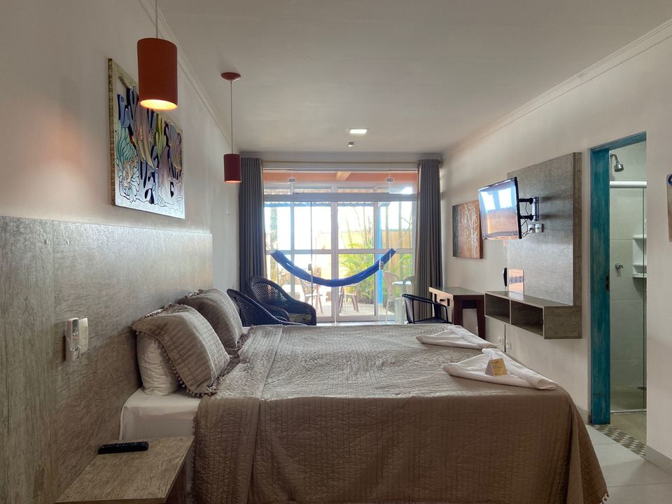 Suíte em Ubatuba com cama de casal, cama de solteiro em outro ambiente e pintura marinha colorida na parede cor de ferrugem. Paredes brancas, cabeceira cinza texturizada, espelho e ar-condicionado branco. Porta aberta para o outro quarto com cama de solteiro e piso de cerâmica clara na Pousada Kaliman