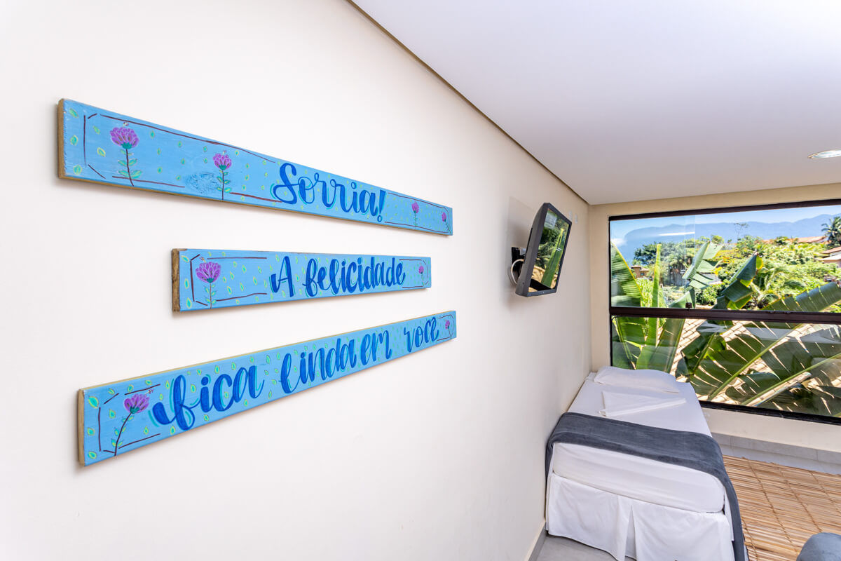 Três placas azuis com uma frase motivacional escrita e ao lado cama de solteiro e janela com vista para o mar e serra de Ilhabela no Eco Baepi Residence