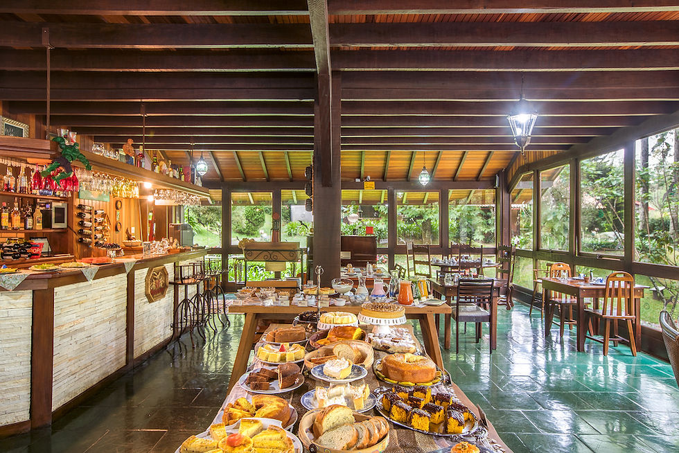 Salão com mesa de café da manhã repleta de pratos, bolos e paes da Pousada Cantos e Contos, localizada em Monte Verde, Minas
