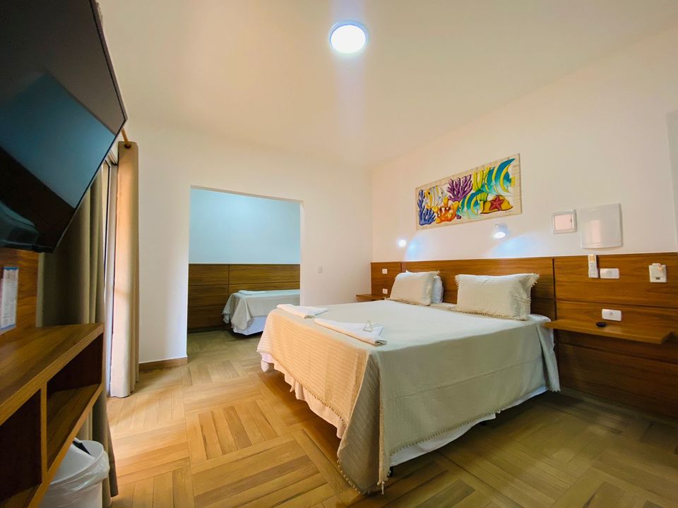 Suíte com cama de casal, cama de solteiro em outro ambiente e pintura marinha colorida na parede. Paredes brancas, cabeceira de madeira clara com arandelas e piso de madeira clara em espinha de peixe. Televisão parcialmente visível e porta com cortina clara na praia de Ubatuba na Pousada Kaliman