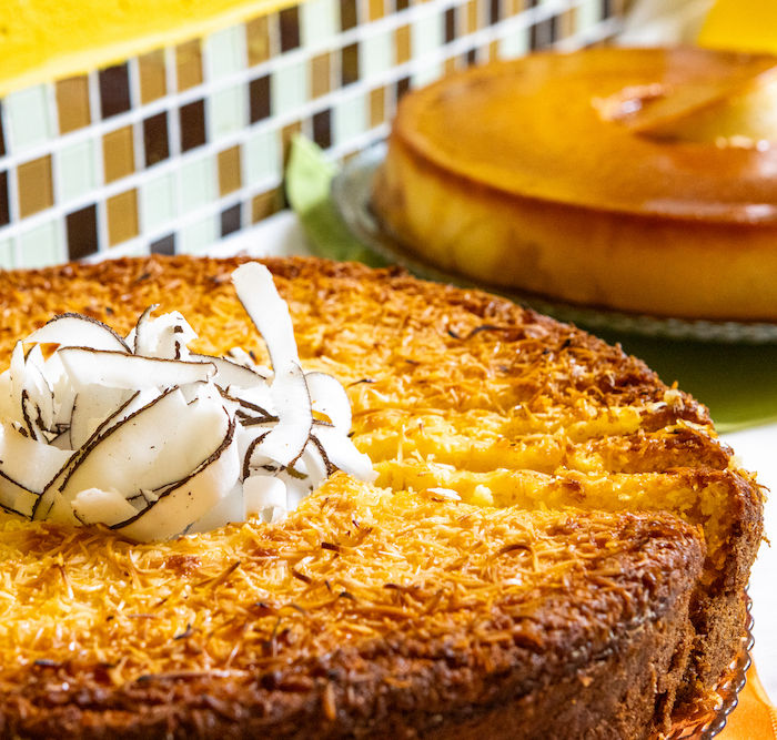 Café da manhã em Ubatuba com um bolo redondo dourado, possivelmente de coco e pêssego com gengibre, é centralizado e decorado com coco. Ao fundo, outro bolo caramelo em prato verde contrasta com a parede de azulejos marrom e bege. A iluminação realça a textura e as cores dos bolos na Pousada Kaliman