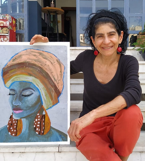 Xuxu Neffa sentada em uma escada sorrindo e mostrando sua pintura de uma mulher com os olhos fechados, pele azul e usando turbante 