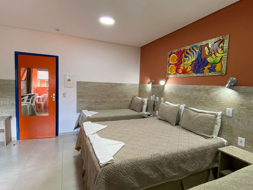 Quarto vibrante na praia de Ubatuba com cama de casal e solteiro em tons de bege. Parede da cabeceira em madeira clara e terracota com quadro de peixes. Porta laranja com espelho à esquerda e balcão funcional com pia e espelho ao lado na Pousada Kaliman