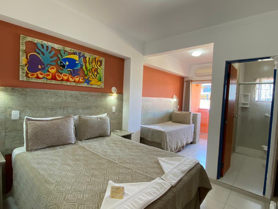 Quarto da Pousada em Ubatuba com cama de casal, sofá-cama em ambiente separado e pintura marinha colorida na parede cor de ferrugem. Paredes brancas, cabeceira cinza texturizada e porta azul para o banheiro parcialmente visível. Sofá-cama contra parede cor de ferrugem com arandela e abertura para área externa iluminada.