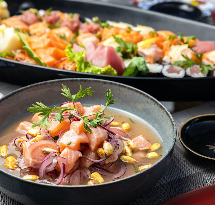 Um close-up de uma tigela cinza contendo ceviche fresco, com cubos de peixe rosa misturados com cebola roxa fatiada e grãos de milho em um caldo claro, guarnecido com folhas de coentro. Ao fundo, um barco preto com mais frutos do mar sugere uma refeição compartilhada no restaurante da Pousada Kaliman