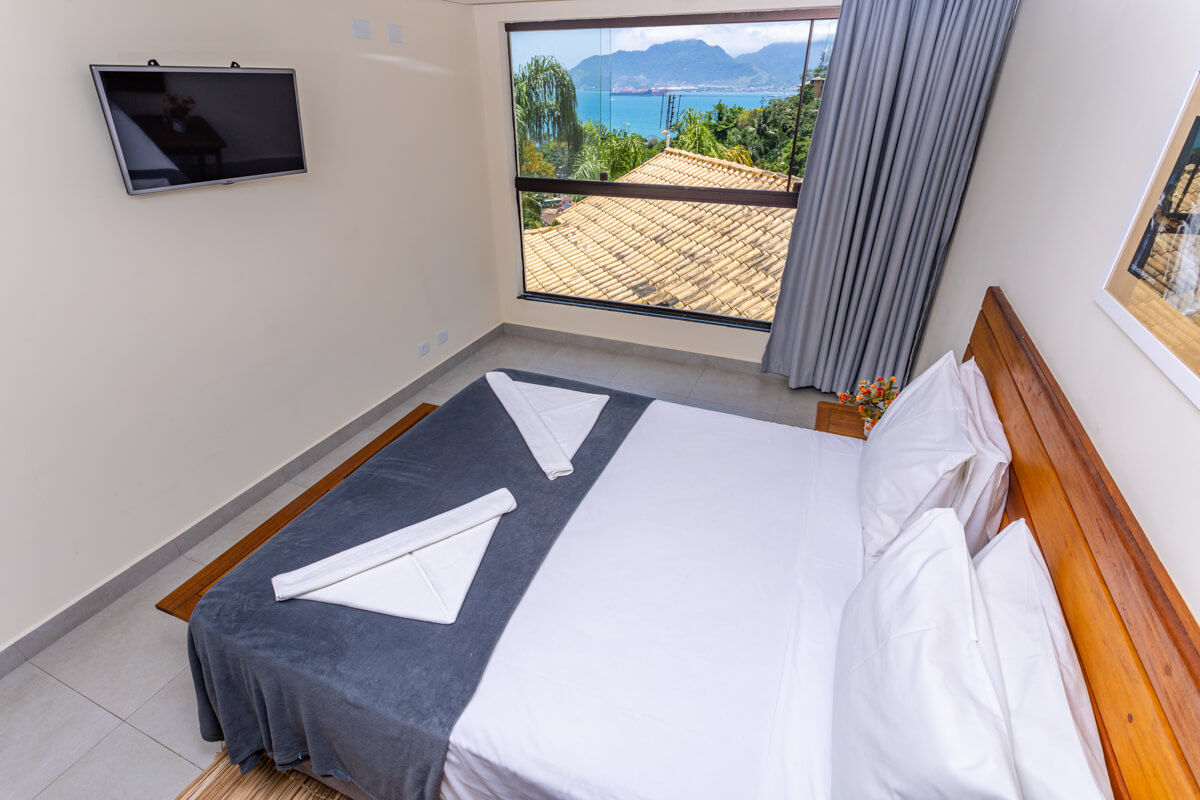 Suíte do Eco Baepi Residence com cama de casal posicionada em frente a televisão e janela com vista para o mar e serra de Ilhabela