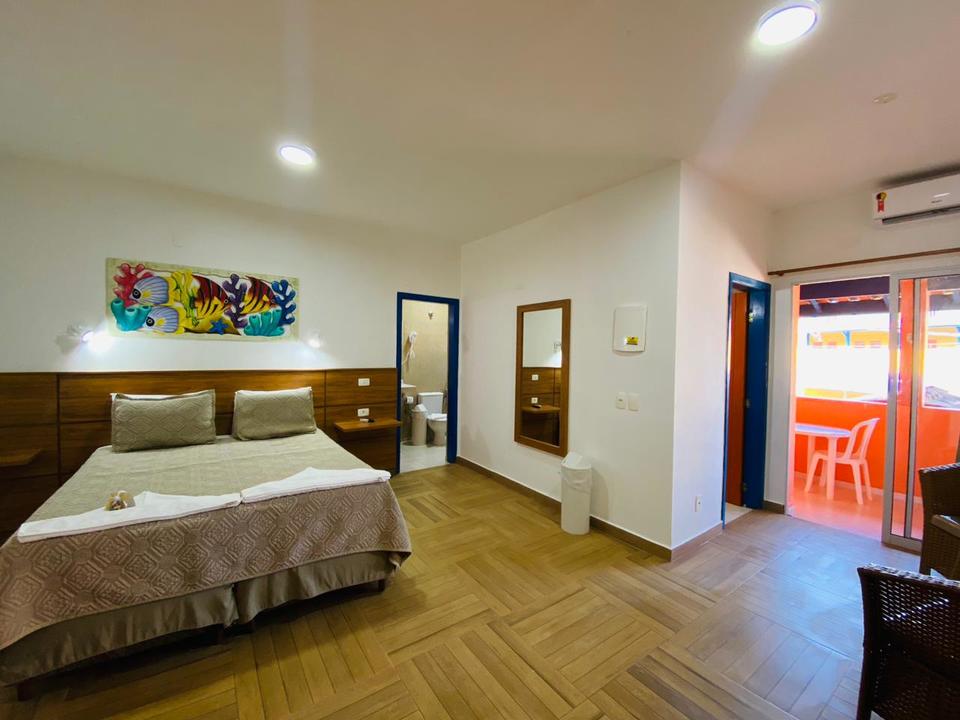 Suíte em Ubatuba com cama de casal, pintura marinha colorida e cabeceira de madeira clara com arandelas. Piso de madeira clara em espinha de peixe e porta azul aberta para banheiro com vaso sanitário branco. Espelho retangular com moldura de madeira e lixeira branca no canto na Pousada Kaliman