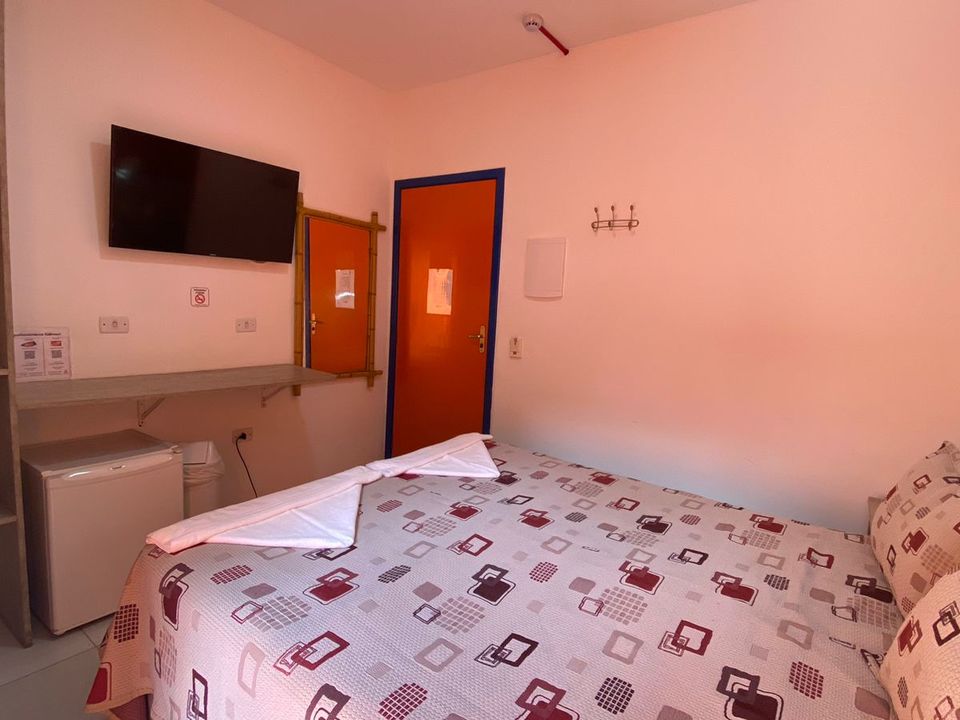 Quarto na Pousada Kaliman em Ubatuba com paredes brancas, duas luminárias pendentes vermelhas e mesa de cabeceira de madeira clara ao lado da cama de casal marrom com travesseiros estampados. Apresenta pintura abstrata sobre cabeceira cinza, porta de vidro para varanda com rede azul e vista para vegetação, TV de tela plana e mesa de madeira com cadeiras.