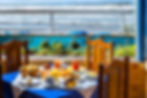 Café da manhã colorido para dois com vista para o mar azul. A mesa está repleta de diversas opções de comida, e a varanda decorada com tema marinho oferece um cenário tropical na Pousada Kaliman na praia de Ubatuba